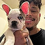 man, smile, curly_hair, nose_ring, puppy, french_bulldog, dog, blue_eye, pink_ears, hand, ring, indoor, wall, person, face, holding, cute, pet, closeup, happy