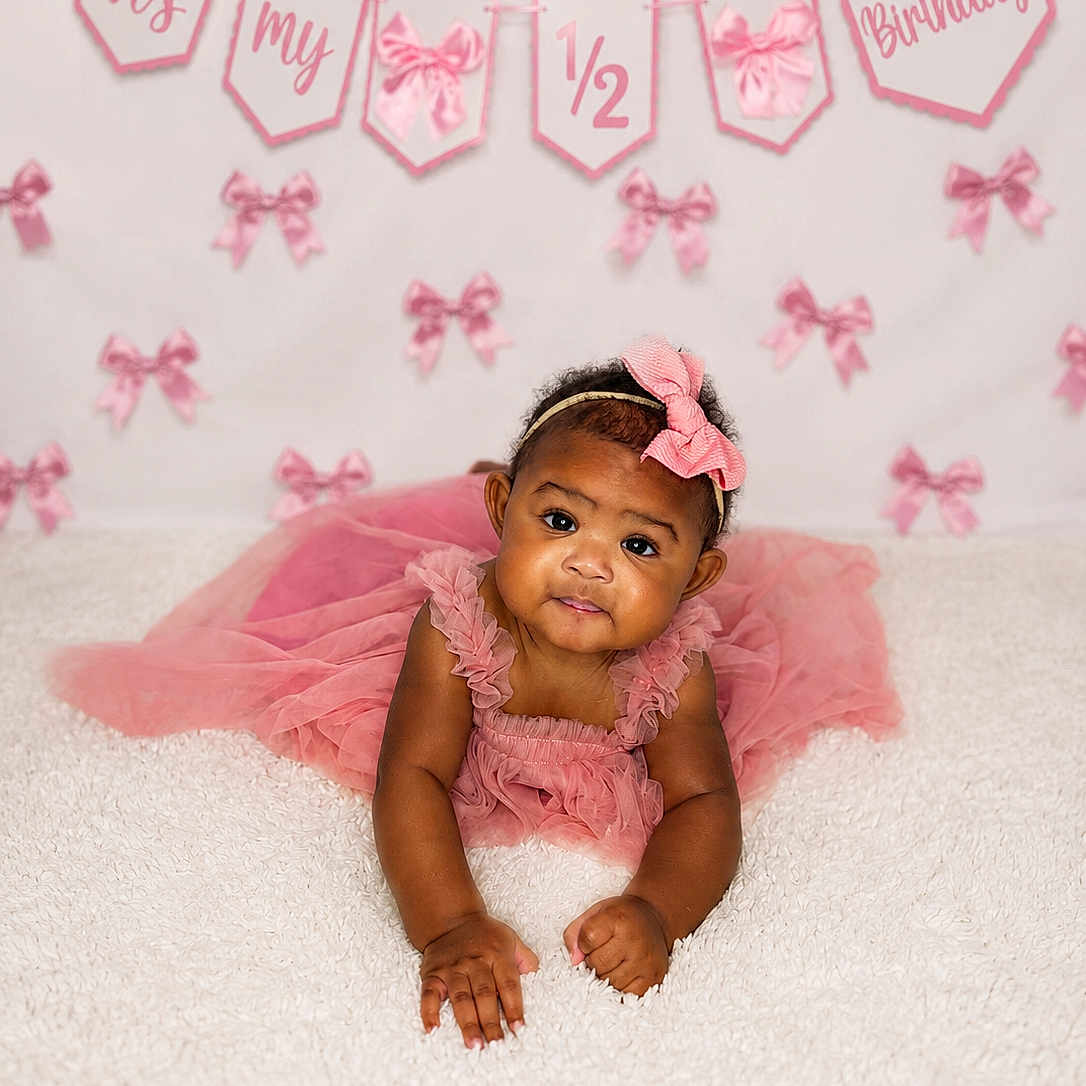 Kabrielle joined the competition — help win amazing prizes! baby, child, infant, pink_dress, headband, bow, carpet, celebration, birthday, half_birthday, banner, decorations, cute, portrait, smiling, happy, lying_down, indoors, soft_texture, curious