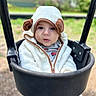 baby, child, swing, playground, outdoor, cute, hooded_jacket, white_clothing, black_swing, face, portrait, infant, winter_clothing, soft_texture, curious, seated, toy_truck_print, nature, blurred_background, daylight