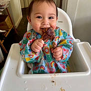 Mae is registered to the contest to win money with this photo: baby, high_chair, bib, food, meat, happy, smiling, messy, indoor, child, face, hands, colorful_clothing, table, feeding, cute, meal, tongue, cheek, dining