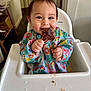 baby, high_chair, bib, food, meat, happy, smiling, messy, indoor, child, face, hands, colorful_clothing, table, feeding, cute, meal, tongue, cheek, dining