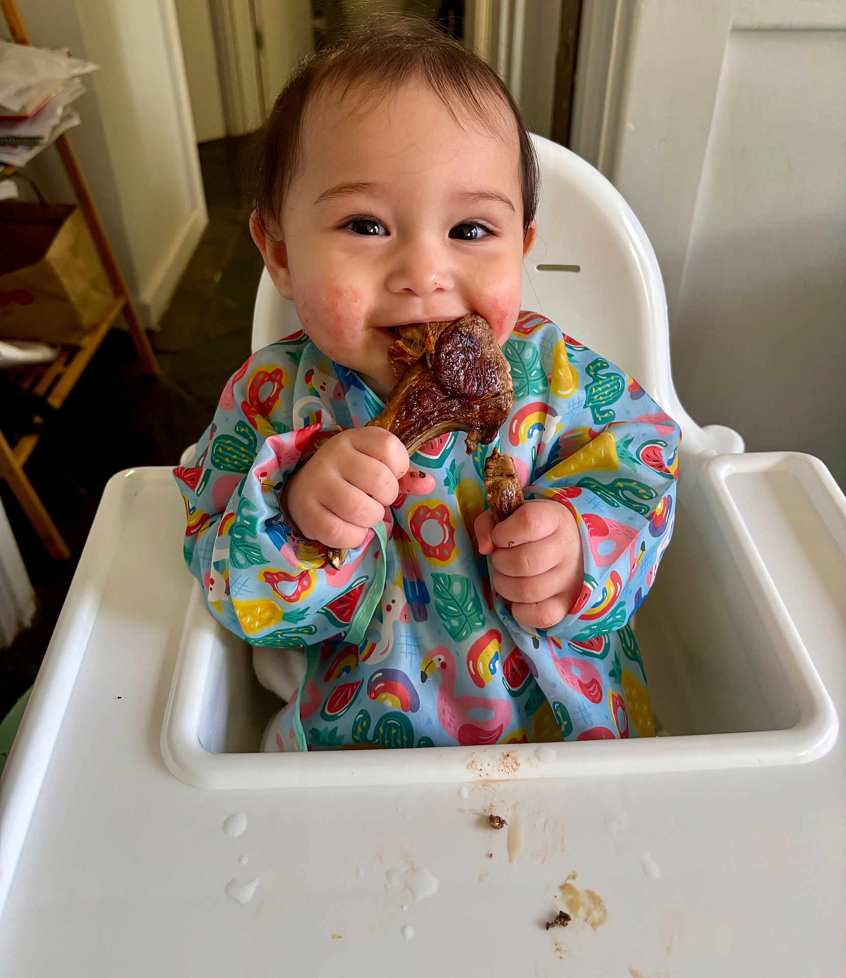 Mae is registered to the contest to win money with this photo: baby, high_chair, bib, food, meat, happy, smiling, messy, indoor, child, face, hands, colorful_clothing, table, feeding, cute, meal, tongue, cheek, dining