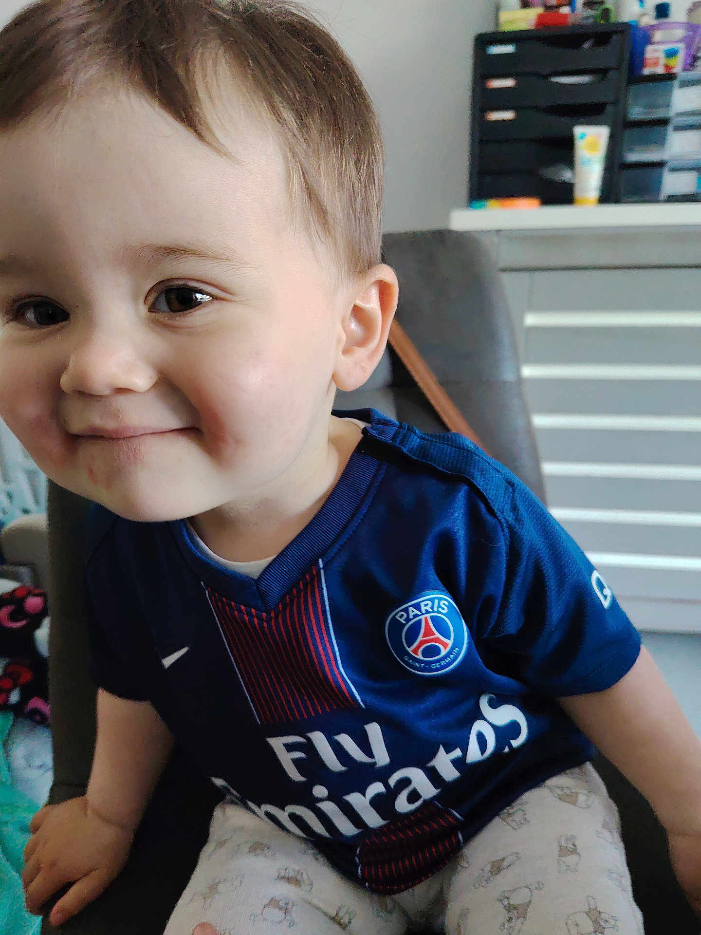 Marlon participe au concours pour gagner de l'argent avec cette photo : toddler, child, smiling, face, blue_jersey, paris_saint_germain, sportswear, indoor, chair, home_office, cabinet, drawer, light, person, cute, happy, short_hair, pants, pattern, casual