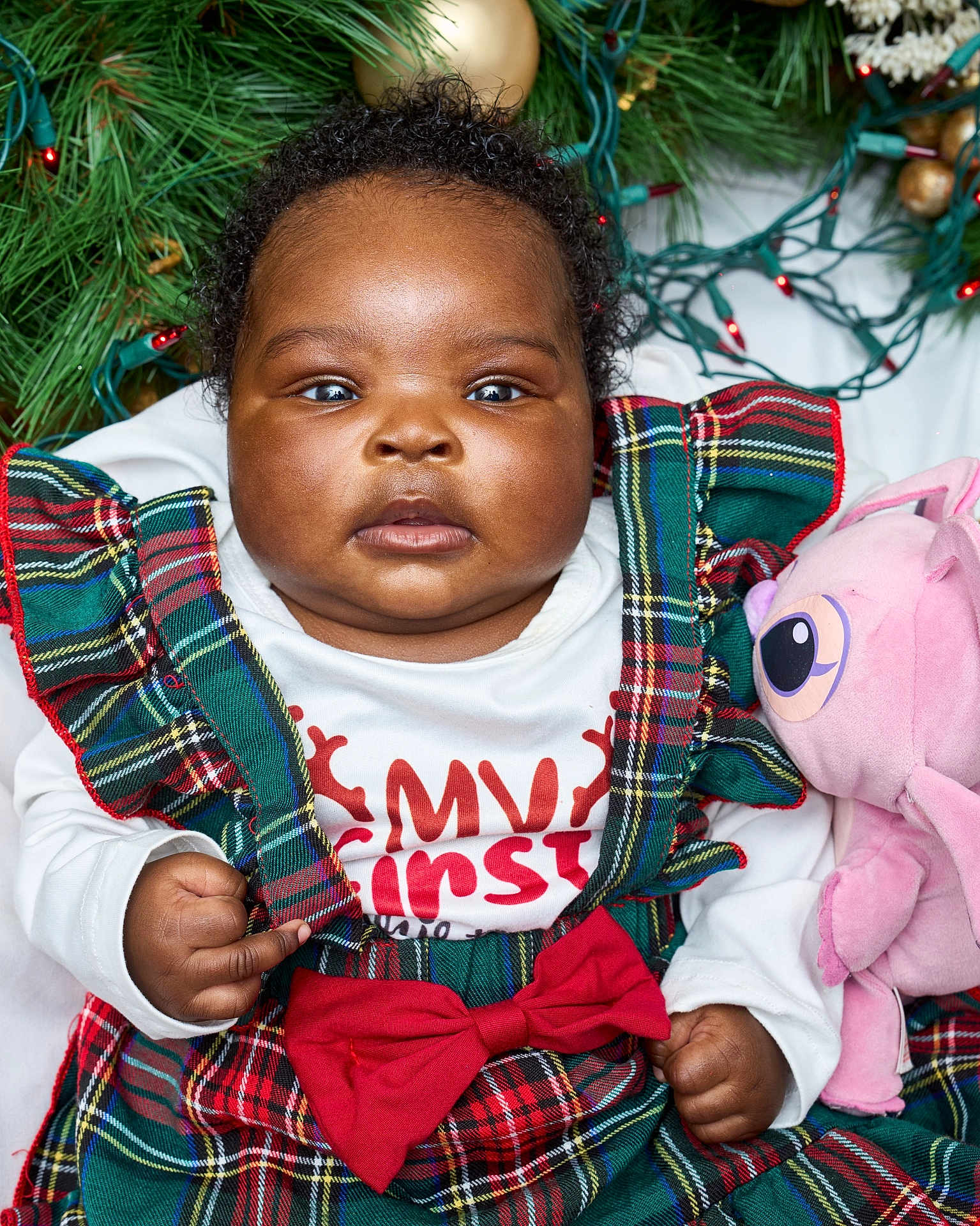 Lauren is registered to the contest to win money with this photo: baby, child, plaid_dress, red_bow, pink_plush_toy, holiday_lights, greenery, curly_hair, festive_clothing, close_up, indoor, cute, portrait, infant, christmas_theme, holiday_season, adorable, smiling, young_child, celebration