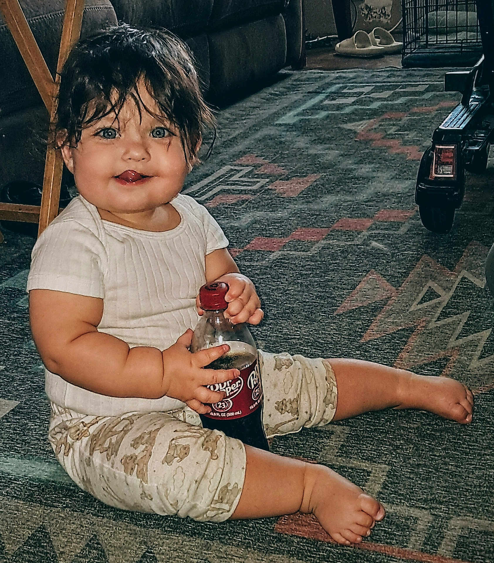 Jesse is registered to the contest to win money with this photo: baby, child, indoor, sitting, carpet, bottle, drink, dr_pepper, clothing, patterned_pants, barefoot, wet_hair, blue_eyes, tongue_out, cute, floor, furniture, slippers, toy_vehicle, happy