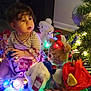 toddler, child, stuffed_animals, christmas_lights, holiday, decorations, indoor, curly_hair, plush_toys, festive, colorful_lights, christmas_tree, toy, child_playing, holiday_decor, candid, cute, warm_lighting, celebration, person