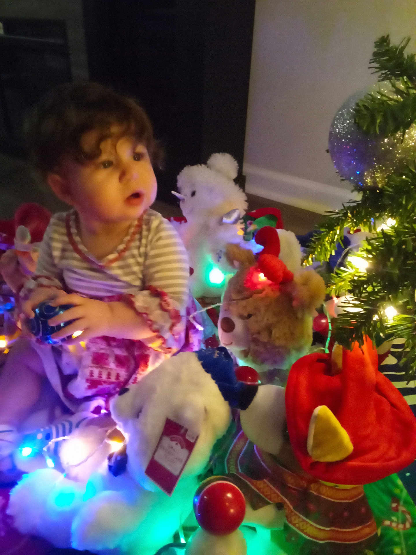 Chloe is registered to the contest to win money with this photo: toddler, child, stuffed_animals, christmas_lights, holiday, decorations, indoor, curly_hair, plush_toys, festive, colorful_lights, christmas_tree, toy, child_playing, holiday_decor, candid, cute, warm_lighting, celebration, person