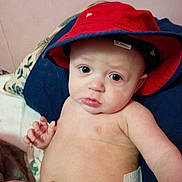Dakota joined the competition — help win amazing prizes! baby, child, hat, red_hat, diaper, indoor, pillow, cushion, face, skin, expression, eyes, hand, sitting, blanket, person, young_child, closeup, portrait, curious