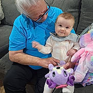 Caroline is registered to the contest to win money with this photo: baby, elderly_person, blue_shirt, glasses, gray_couch, plush_toy, purple_toy, colorful_toy, smile, indoor, person, seated, happy, family, cute, child, hand_holding, cozy, casual_clothing, wooden_floor