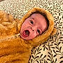 baby, infant, face, mouth_open, eyes, fuzzy_clothing, hood, ears, blanket, pattern, leaf_pattern, bed, soft_texture, cute, lying_down, expression, child, warm_clothing, indoor, closeup