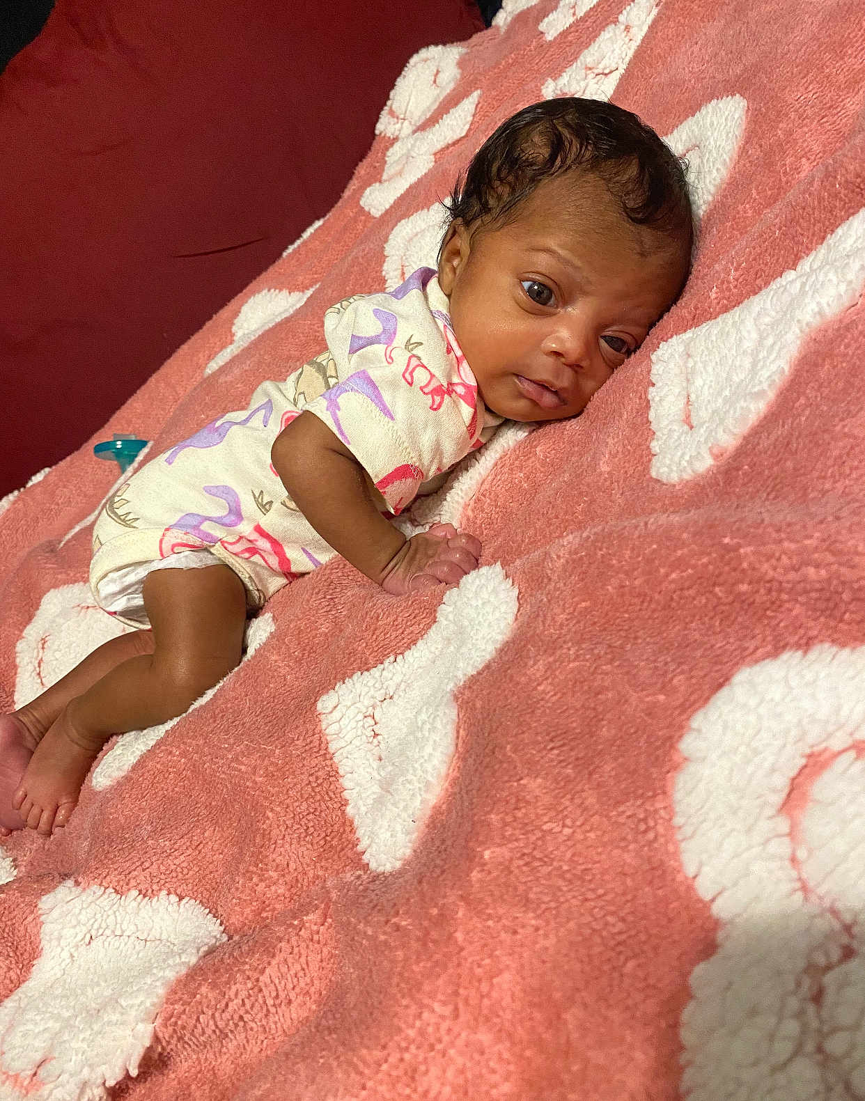Kyari is registered to the contest to win money with this photo: baby, infant, blanket, pink, white, onesie, dinosaur_pattern, lying_down, curious, face, skin, foot, hand, sleepwear, indoors, soft_texture, cute, child, newborn, resting