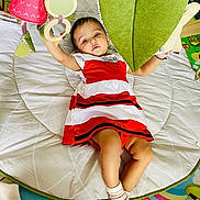 Veda joined the competition — help win amazing prizes! baby, child, red_and_white_dress, play_mat, toys, hanging_toys, green_cushion, socks, indoor, floor_mat, colorful, infant, person, cute, reaching, lying_down, soft_toys, play_area, young_child, comfortable