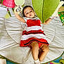 baby, child, red_and_white_dress, play_mat, toys, hanging_toys, green_cushion, socks, indoor, floor_mat, colorful, infant, person, cute, reaching, lying_down, soft_toys, play_area, young_child, comfortable