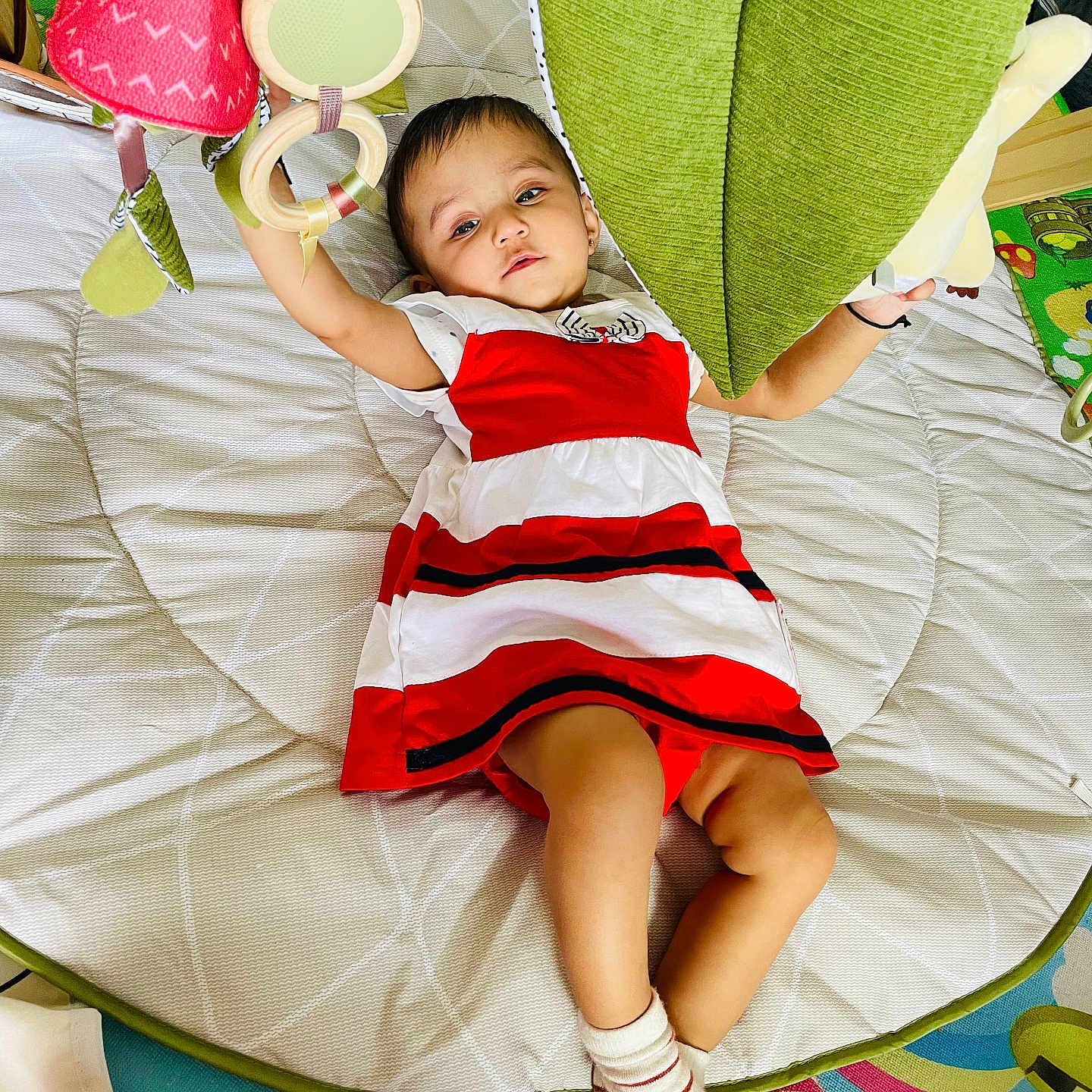 Veda joined the competition — help win amazing prizes! baby, child, colorful, comfortable, cute, floor_mat, green_cushion, hanging_toys, indoor, infant, lying_down, person, play_area, play_mat, reaching, red_and_white_dress, socks, soft_toys, toys, young_child