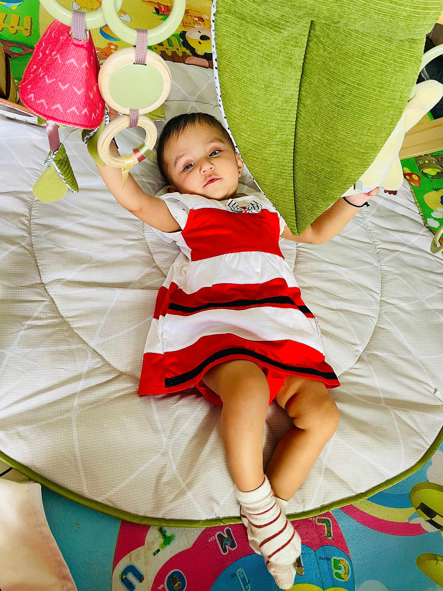 Veda joined the competition — help win amazing prizes! baby, child, red_and_white_dress, play_mat, toys, hanging_toys, green_cushion, socks, indoor, floor_mat, colorful, infant, person, cute, reaching, lying_down, soft_toys, play_area, young_child, comfortable
