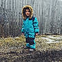 child, toddler, snowsuit, blue_clothing, hood, fur_trim, outdoor, winter, muddy_ground, grass, wooden_fence, smiling, standing, cold_weather, boots, person, nature, portrait, casual, daytime
