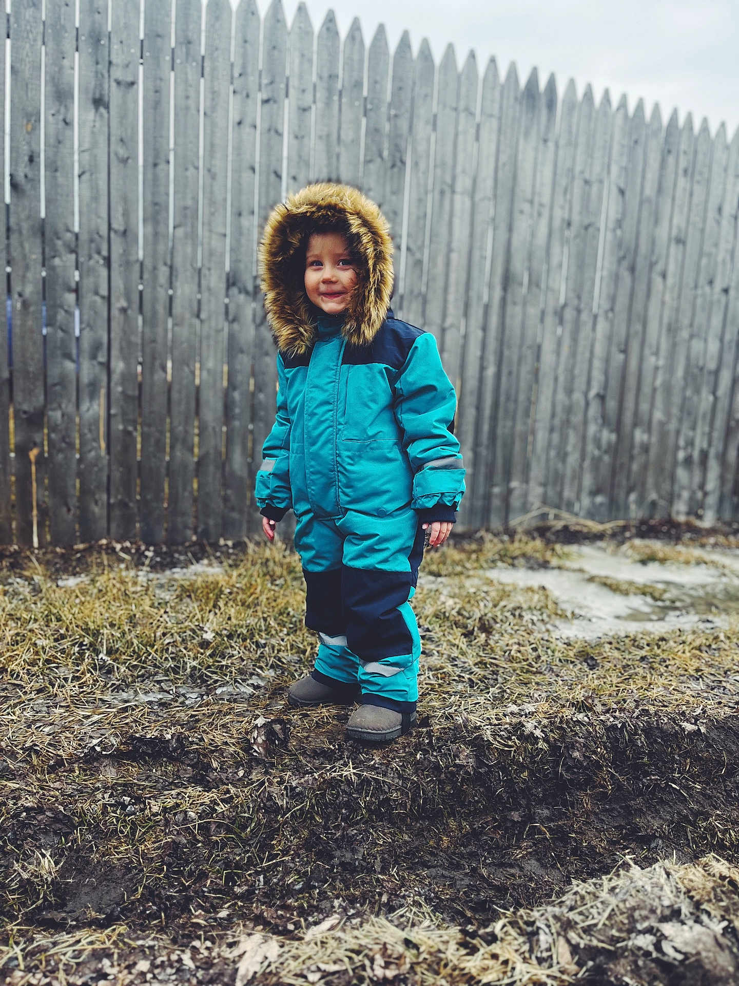 Willow is registered to the contest to win money with this photo: child, toddler, snowsuit, blue_clothing, hood, fur_trim, outdoor, winter, muddy_ground, grass, wooden_fence, smiling, standing, cold_weather, boots, person, nature, portrait, casual, daytime