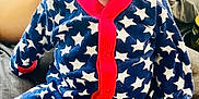 Stetson is registered to the contest to win money with this photo: baby, infant, onesie, stars_pattern, red_trim, hand, face, blue, clothing, indoor, person, newborn, cute, seated, soft_surface, curious_expression, human, small, cozy, support
