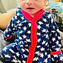 baby, infant, onesie, stars_pattern, red_trim, hand, face, blue, clothing, indoor, person, newborn, cute, seated, soft_surface, curious_expression, human, small, cozy, support