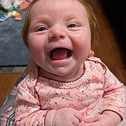Peyton is registered to the contest to win money with this photo: baby, smiling, happy, infant, child, face, portrait, pink_clothing, floral_pattern, hand, tattoo, skin, indoor, person, cute, young, closeup, joy, expression, holding