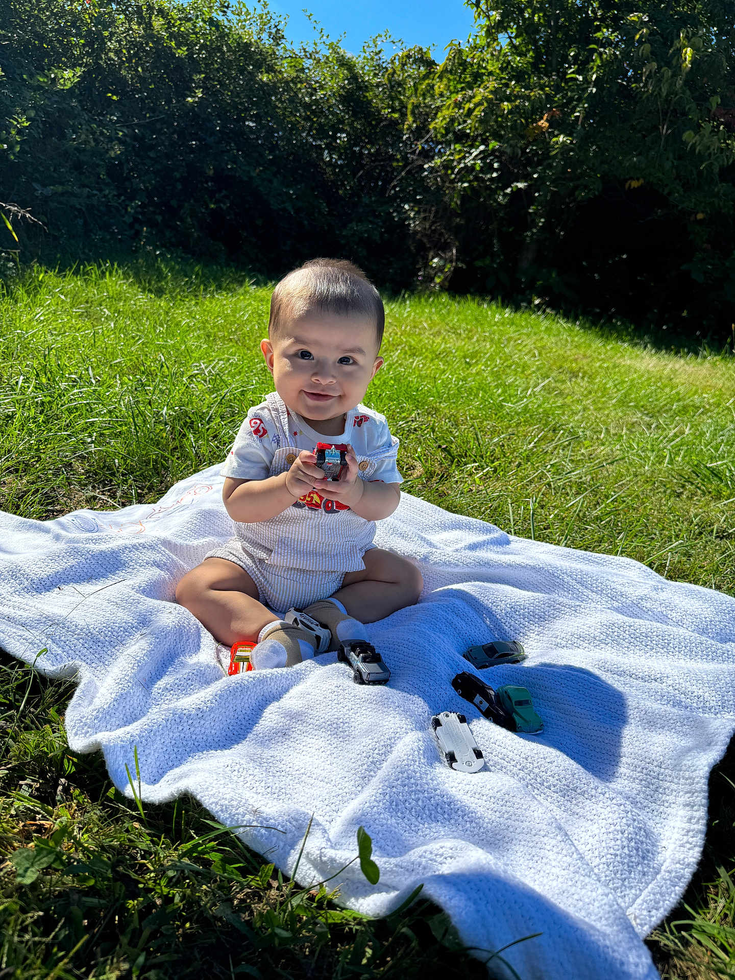 Leo joined the competition — help win amazing prizes! baby, toy_car, blanket, grass, outdoor, greenery, sunlight, smile, child, nature, play, sitting, happy, shorts, shirt, footwear, shadow, leaf, daytime, toy