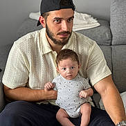Raphaël participe au concours pour gagner de l'argent avec cette photo : man, baby, child, person, indoor, couch, seated, cap, beard, knit_shirt, onesie, curious, holding, living_room, watching, casual, home, furniture, relaxed, portrait