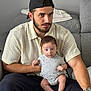 man, baby, child, person, indoor, couch, seated, cap, beard, knit_shirt, onesie, curious, holding, living_room, watching, casual, home, furniture, relaxed, portrait