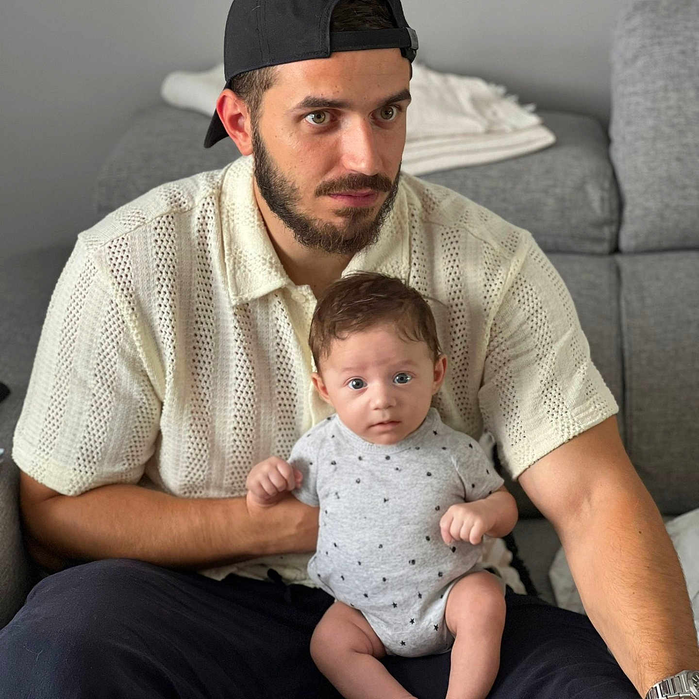 Raphaël participe au concours pour gagner de l'argent avec cette photo : baby, beard, cap, casual, child, couch, curious, furniture, holding, home, indoor, knit_shirt, living_room, man, onesie, person, portrait, relaxed, seated, watching
