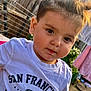 toddler, child, face, hair, white_tshirt, text_on_clothing, outdoor, fence, laundry, curious, portrait, young_child, casual_clothing, daylight, expression, person, cute, closeup, garden, playful