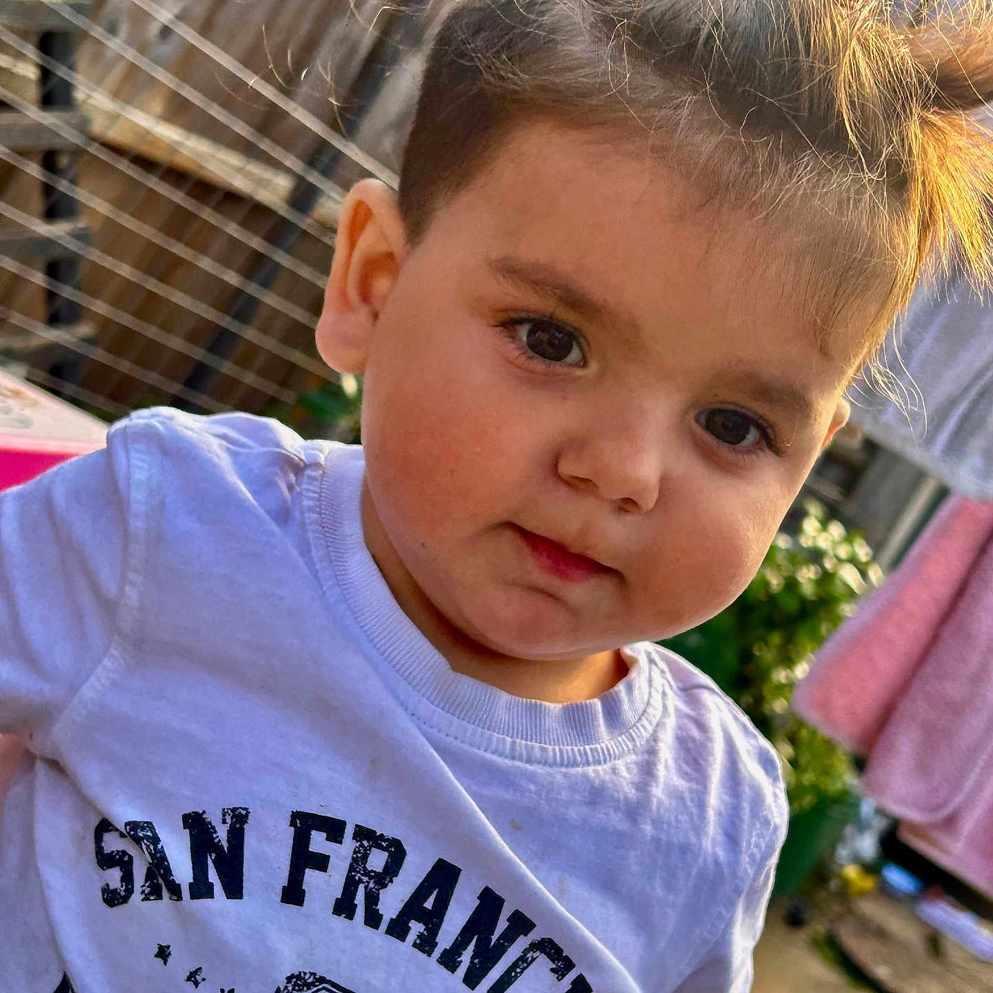 Kasian is registered to the contest to win money with this photo: casual_clothing, child, closeup, curious, cute, daylight, expression, face, fence, garden, hair, laundry, outdoor, person, playful, portrait, text_on_clothing, toddler, white_tshirt, young_child