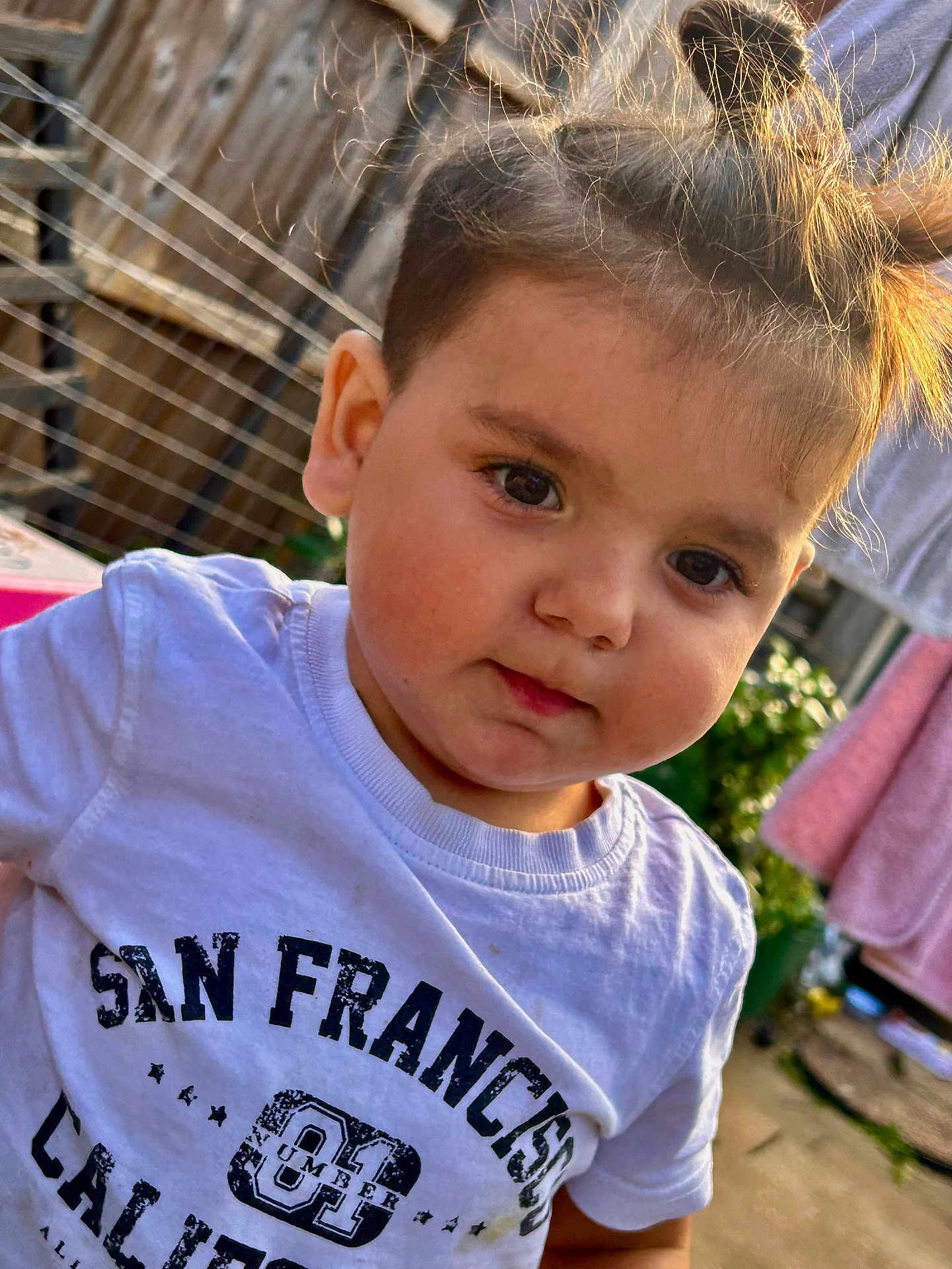Kasian is registered to the contest to win money with this photo: toddler, child, face, hair, white_tshirt, text_on_clothing, outdoor, fence, laundry, curious, portrait, young_child, casual_clothing, daylight, expression, person, cute, closeup, garden, playful