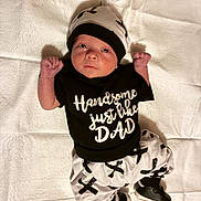 Xavier is registered to the contest to win money with this photo: newborn, baby, infant, child, portrait, lying_down, quilt, blanket, hat, beanie, black_clothing, white_clothing, onesie, pants, shoes, raised_fists, serious_expression, indoor, fashion, text_on_clothing