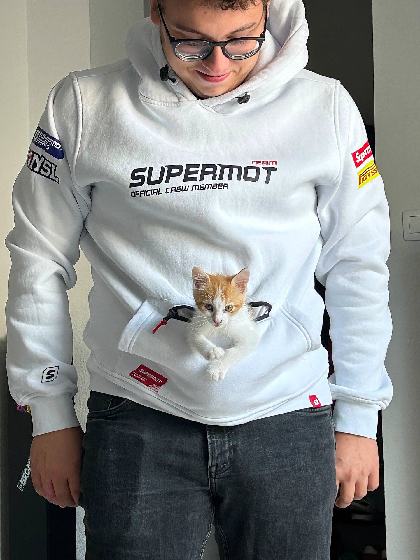 affection, casual_clothing, cat, cozy, cute, glasses, hoodie, indoor, jeans, kitten, looking_down, man, orange_and_white_cat, person, pet, pocket, smiling, standing, white_hoodie, young_adult