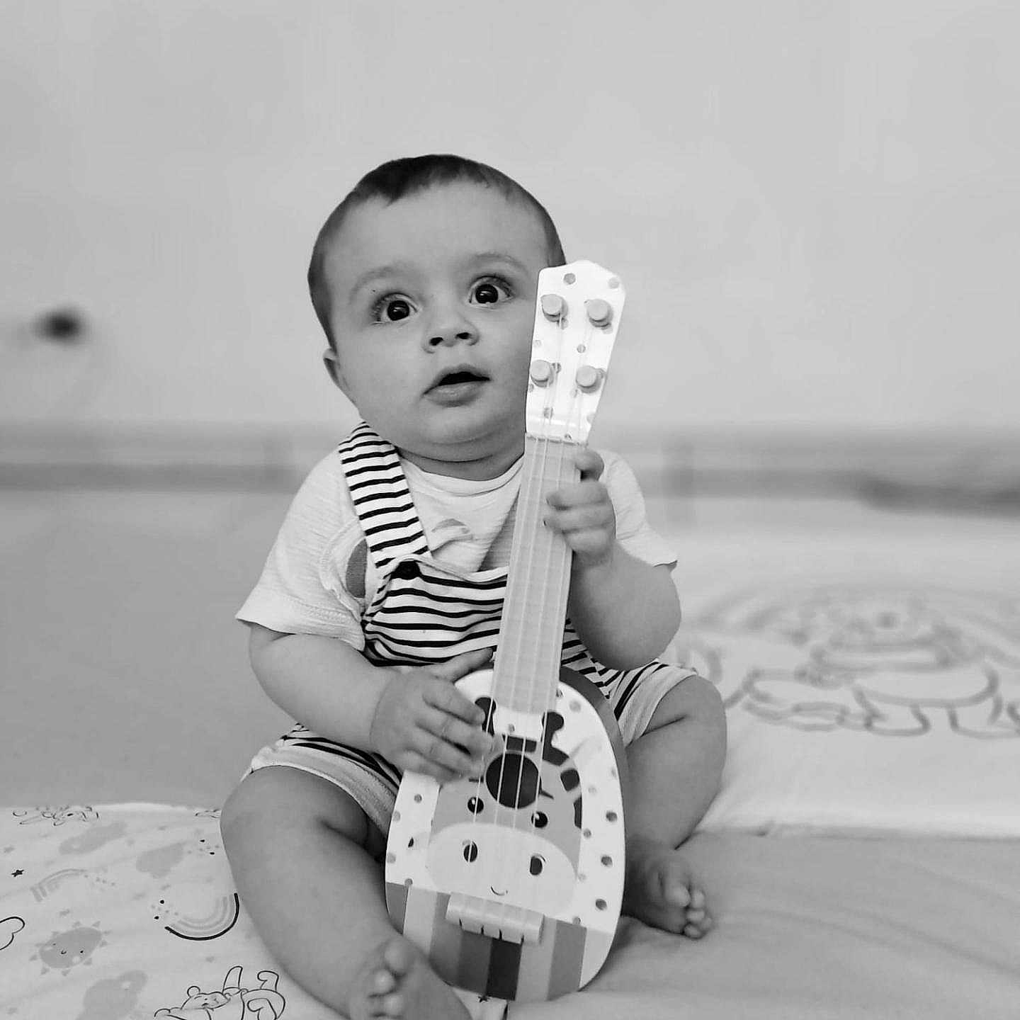 Leyan participe au concours pour gagner de l'argent avec cette photo : baby, black_and_white, blanket, child, curious, cute, guitar, holding, indoor, infant, musical_instrument, patterned_blanket, playful, portrait, sitting, small_feet, striped_clothing, toy, wide_eyes, young