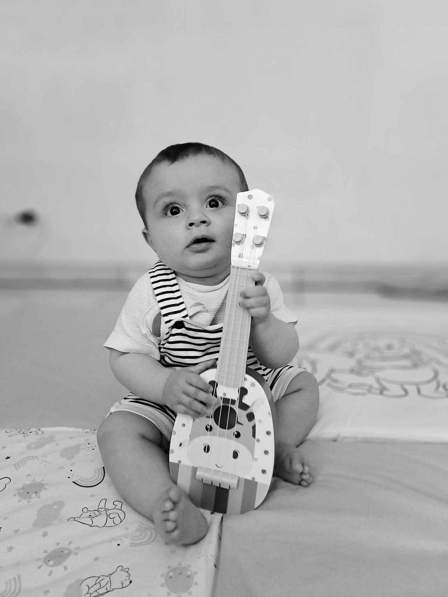 Leyan participe au concours pour gagner de l'argent avec cette photo : baby, child, toy, guitar, musical_instrument, striped_clothing, sitting, indoor, blanket, patterned_blanket, cute, curious, black_and_white, portrait, infant, small_feet, holding, playful, wide_eyes, young