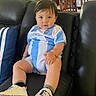 Logan is registered to the contest to win money with this photo: baby, infant, child, couch, leather, blue_and_white, argentina, soccer_jersey, pillow, footwear, socks, indoors, furniture, curly_hair, cute, portrait, seated, home, casual, looking_at_camera