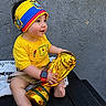 baby, child, trophy, golden_trophy, yellow_shirt, headband, ecuador_flag, bench, barefoot, happy, smiling, outdoor, curious, shorts, wristwatch, portrait, sitting, cute, infant, playful