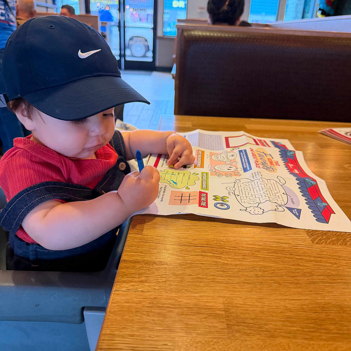 Logan is registered to the contest to win money with this photo: toddler, child, hat, cap, red_shirt, high_chair, coloring, crayon, placemat, table, restaurant, indoor, person, seated, wooden_table, activity_sheet, lighting, casual, diner, focused