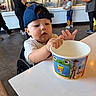 toddler, child, cup, toy_story, blue_cap, frozen_yogurt_shop, table, high_chair, people, background, floor, clothing, indoor, curious, hand, face, portrait, young, food_container, waiting