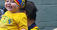 Logan is registered to the contest to win money with this photo: baby, child, smiling, headband, yellow_shirt, adult, holding, outdoor, wall, poster, colorful, person, happy, face, hand, clothing, portrait, people, head, casual
