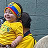 baby, child, smiling, headband, yellow_shirt, adult, holding, outdoor, wall, poster, colorful, person, happy, face, hand, clothing, portrait, people, head, casual