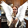 person, cat, kitchen, wooden_cabinets, stove, angel_wings, glasses, gray_hoodie, flooring, countertop, animal, pet, indoor, surprised_expression, holding, digital_effect, white_wings, domestic_cat, home, funny