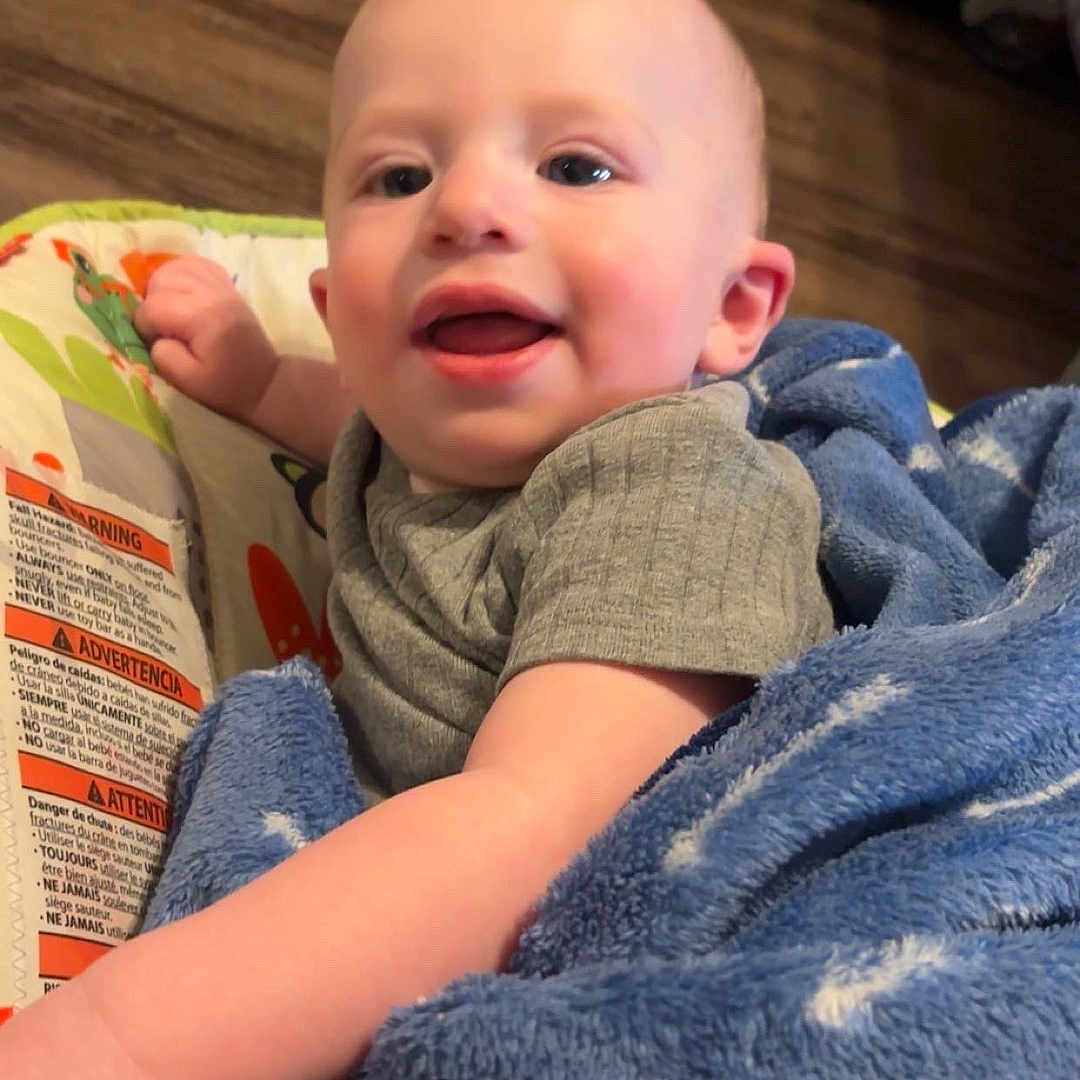 Clayton joined the competition — help win amazing prizes! baby, child, blanket, blue_blanket, baby_seat, indoor, happy, smiling, infant, person, floor, wooden_floor, furniture, cozy, cute, portrait, clothing, hand, face, young