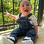 baby, child, smiling, denim_overalls, white_tshirt, nike_logo, sneakers, outdoor, sunlight, greenery, metal_railing, happy, cute, portrait, sitting, infant, casual_clothing, daylight, concrete_floor, expressive_face
