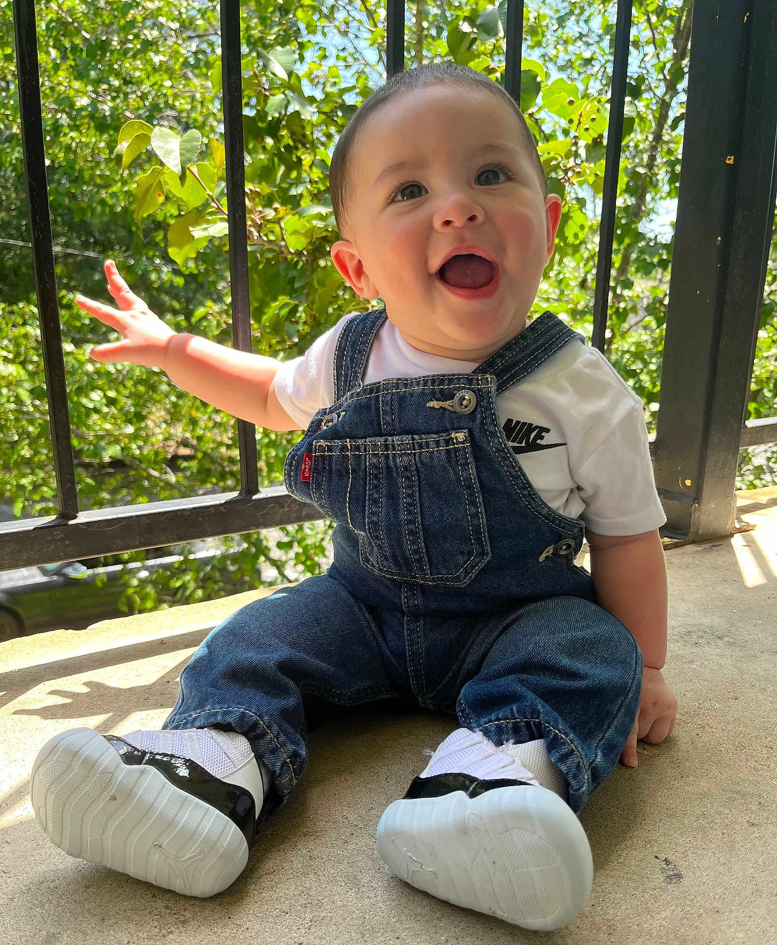 Eithan Loaiza is registered to the contest to win money with this photo: baby, child, smiling, denim_overalls, white_tshirt, nike_logo, sneakers, outdoor, sunlight, greenery, metal_railing, happy, cute, portrait, sitting, infant, casual_clothing, daylight, concrete_floor, expressive_face