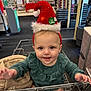 Abigail joined the competition — help win amazing prizes! baby, bag, child, curious, cute, eyes, face, festive, green_clothing, hand, headwear, holiday, indoor, metal, person, retail, santa_hat, shopping_cart, smiling, store