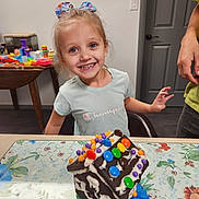 Ariyanna is registered to the contest to win money with this photo: child, girl, smile, happy, indoor, table, gingerbread_house, candy, decorated, bow, hair_accessory, person, adult_hand, clothing, tshirt, chair, door, wall, toy, tray