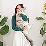 braids, casual_clothing, cat, cozy, domestic, fluffy_cat, greenery, holding, home, indoor, long_hair, pet, plant, portrait, relaxed, skirt, smile, sweater, white_wall, woman