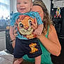 baby, child, smiling, person, holding, kitchen, table, clothing, shorts, tshirt, cartoon, lion_king, adult, hair, window, cupboard, fridge, floor, hands, indoor