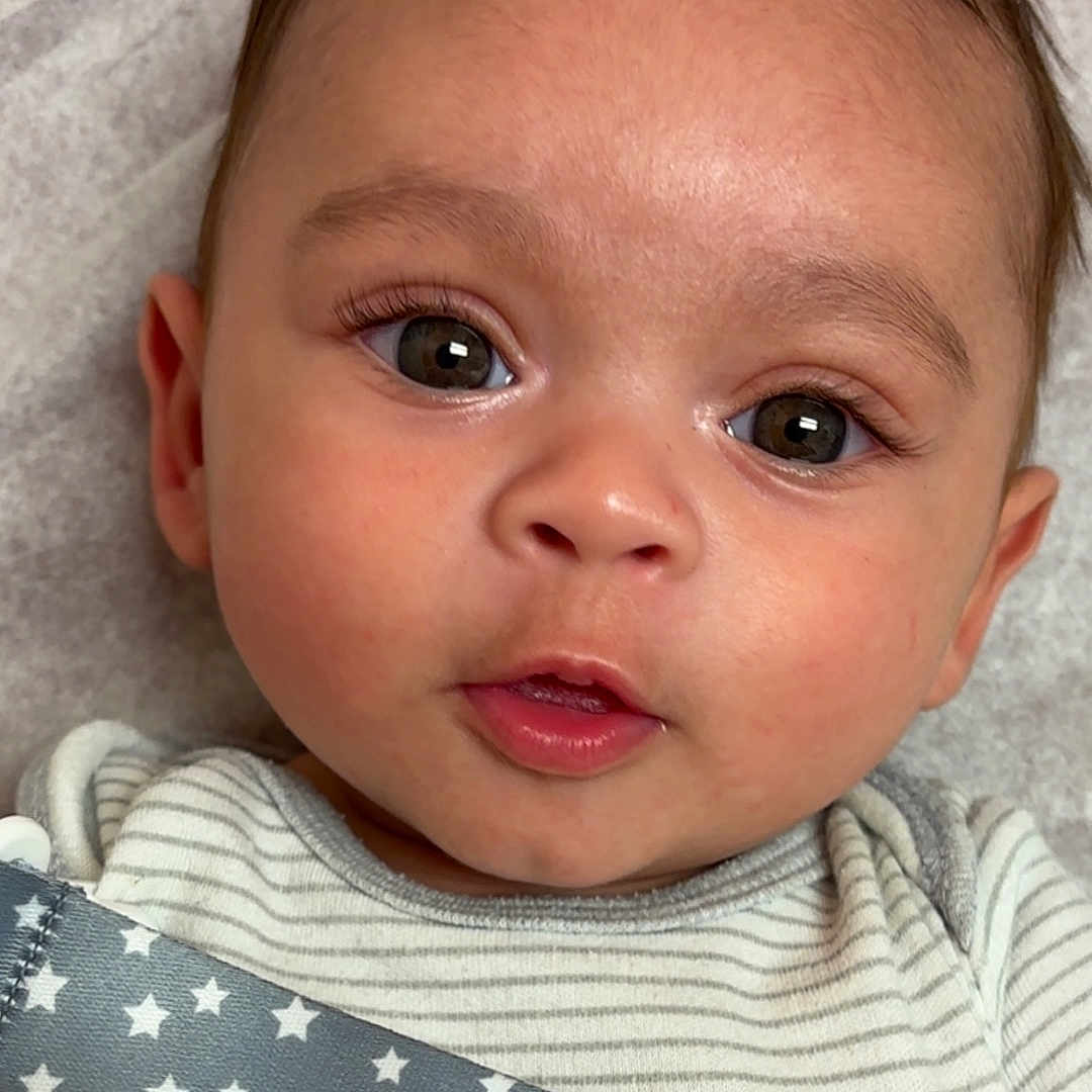 Yadiel is registered to the contest to win money with this photo: baby, cheeks, closeup, cute, expression, eyes, face, head, indoors, infant, lying_down, mouth, onesie, person, portrait, skin, star_pattern, strap, striped_clothing, young_child